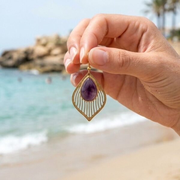 Boucles d'oreilles gouttes Amaya en améthyste naturelle et monture dorée ciselée, présentées à la main devant la mer.