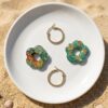 Composition de bijoux : une fleur en papier Washi motif floral, une fleur en résine émeraude avec paillettes dorées, et une paire de créoles dorées hypoallergéniques, le tout sur une petite assiette en céramique.