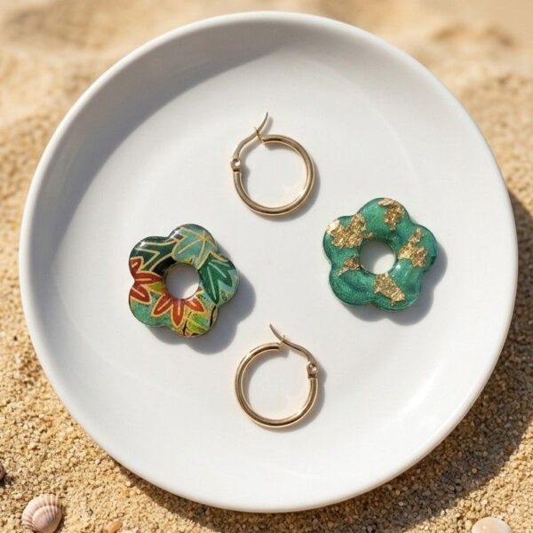 Composition de bijoux : une fleur en papier Washi motif floral, une fleur en résine émeraude avec paillettes dorées, et une paire de créoles dorées hypoallergéniques, le tout sur une petite assiette en céramique.
