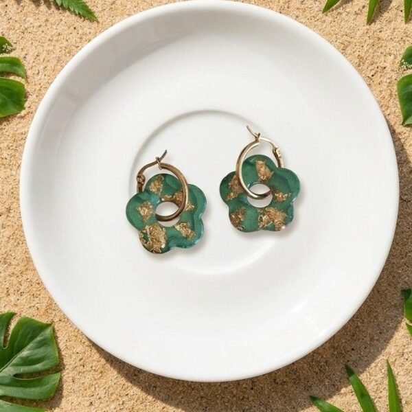 Gros plan d'une paire de boucles d'oreilles créoles dorées avec des pendentifs fleurs en résine verte et pétales dorées