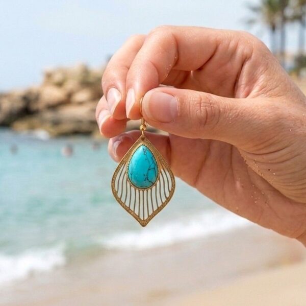 Amaya en Turquoise naturelle et monture dorée ciselée, présentées à la main devant la mer.
